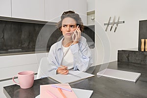 Portrait of woman working from home, calling someone on mobile phone, looking confused while holding pen and trying to