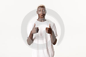 Portrait of wondered and impressed african-american blond man, showing thumbs-up fascinated, standing white background