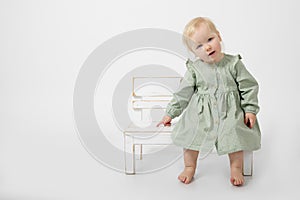 Portrait of wonderful little blond barefoot girl with blue eyes sit on white wooden little bench on white background.