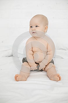 Portrait of wonderful smiling grey-eyed plump cherubic baby infant toddler wearing grey pants sitting on white bed.