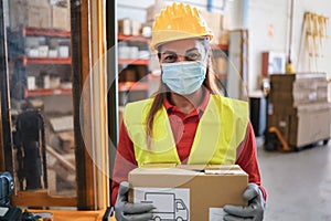 Portrait of worker woman holding cardboard box inside warehouse while wearing safety mask - Focus on face
