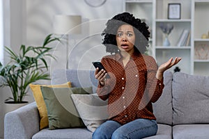 Portrait of worried African American woman sitting on couch at home with phone in hands and holding head in shock