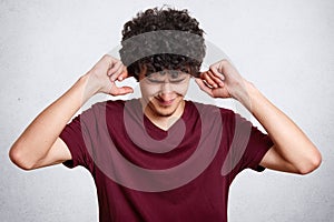 Portrait of worried handsome bearded man in casual maroon t shirt, standing with closed eyes and putting fingers in ears, does not