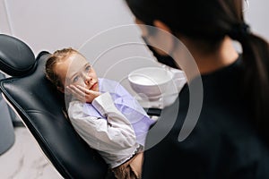 Portrait of worried looking child girl lying on dental chair in front of doctor complaining of toothache and holding