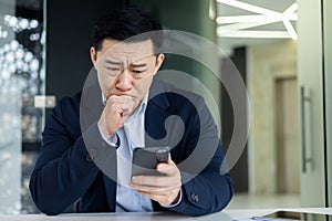 Portrait of a worried young Asian man sitting in the office at the table, holding his head with his hand, looking sadly
