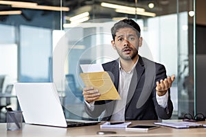 Portrait of a worried young Indian man working in the office, sitting at the desk and holding an open envelope with a