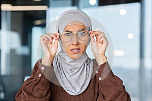 Portrait of worried young muslim woman in hijab sitting in office and looking surprised at camera holding glasses with