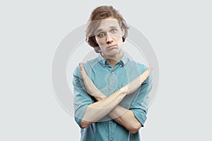 Portrait of worry handsome long haired blonde young man in blue casual shirt standing with x sign hands and looking at camera