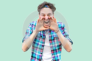 Portrait of yelling young man in casual blue checkered shirt and headband standing with hands on his face, shouting or screaming