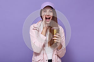 Portrait of young adorable woman, wearing white shirt, pink jacket and cap, has long hair, holding thermomug of coffee, looking at