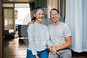 Portrait of young adult Asian couple embracing together with home interior in background. 30s happy mature husband and