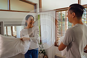 Portrait of young adult Asian couple playing pillow fight in bedroom interior scene. 30s mature husband and wife smiling