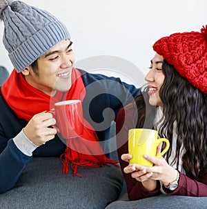 Portrait of young adult Asian lover couple in sweatshirt, scarf, knitted hat relaxing on a couch while holding and drinking hot