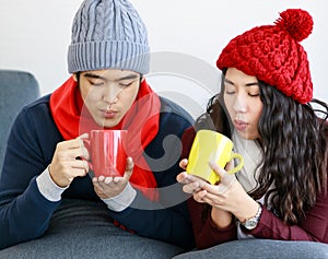 Portrait of young adult Asian lover couple in sweatshirt, scarf, knitted hat relaxing on a couch while holding and drinking hot