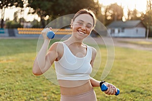 Portrait of young adult attractive smiling woman, being happy to workout in stadium outdoor, holding blue dumbbells in hands,