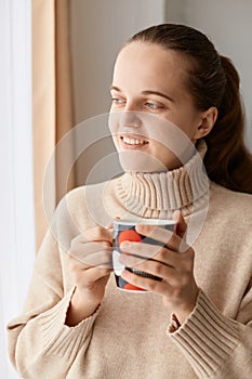 Portrait of young adult attractive woman wearing beige sweater holding cup of coffee or tea, looking away at window, dreaming