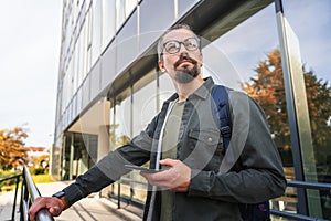 Portrait of young adult caucasian attractive hipster male wearing eyeglasses using smart phone while standing near