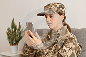 Portrait of young adult Caucasian woman soldier wearing camouflage uniform and cap, sitting on sofa an using smart phone, military