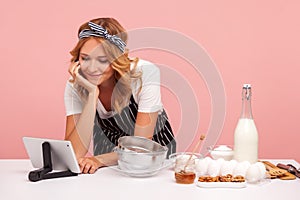 Portrait of young adult female chef wearing pron using tablet for learning new recipe of pastry, looking at digital gadget and