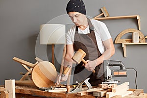 Portrait of young adult man carpenter wearing white t-shirt, black cap and brown apron, craftsman carve with a gouge in the hands