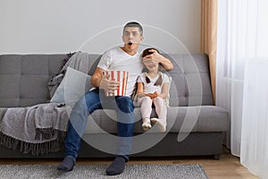 Portrait of young adult man watching horror movie at home with his daughter, covering eyes to his kid, sitting on sofa and holding