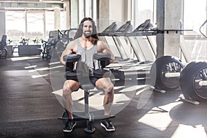 Portrait of young adult muscular built handsome athlete with long curly hair working out in a gym, sitting on a weightlifting