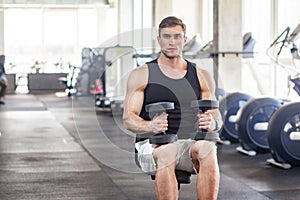 Portrait of young adult muscular built handsome athlete working out in a gym, sitting on a weightlifting machine and holding two