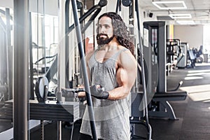 Portrait of young adult sport athlete man with long curly hair training at gym alone, standing and lifting weights in the gym,