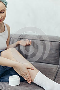 Portrait of young adult woman applying body lotion for skin on legs while sitting on couch at home