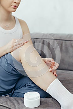 Portrait of young adult woman applying body lotion for skin on legs while sitting on couch at home