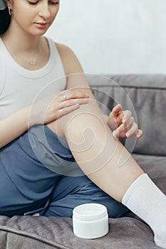 Portrait of young adult woman applying body lotion for skin on legs while sitting on couch at home