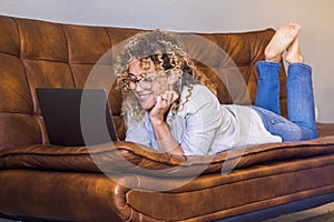 Portrait of young adult woman enjoying indoor relax leisure activity surfing the web on a laptop laying down on the sofa at home.