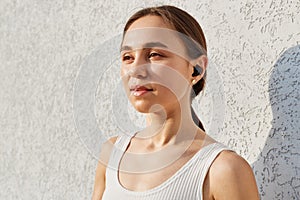 Portrait of young adult woman with pleasant appearance with airpods looking away with dreamy expression, wearing white sporty top