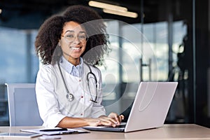 Portrait of a young African American female doctor in a white coat sitting at a table and working at a laptop, looking