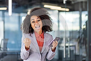 Portrait of a young African-American woman in a suit standing in the office, holding a tablet and showing a success and