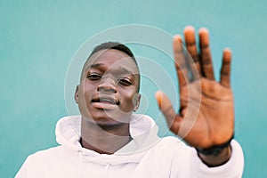 Portrait of young african black man against a green wall with white sweatshirt doing stop sign with is hand. Stop racism protest c