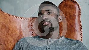 Portrait of young African male sitting in leather chair, thinking about something and smiling. Man looks thoughtfully.