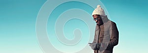 Portrait of young african man with smartphone wearing winter hat, jacket on blue sky background, blank copy space for advertising