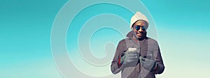 Portrait of young african man with smartphone wearing winter hat, jacket on blue sky background, blank copy space for advertising