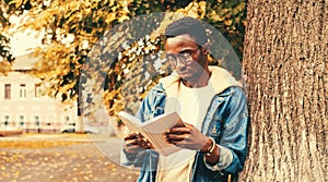 Portrait of young african man student reading a book wearing eyeglasses in autumn city park