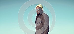 Portrait of young african man wearing sunglasses, winter hat, jacket on sky background
