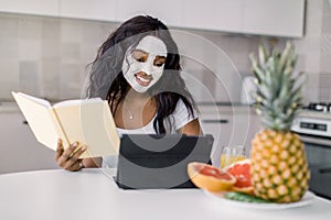 Portrait of young African woman with mud facial mask on her face, sitting at the table with fresh fruits in modern