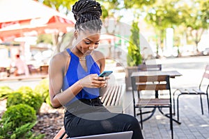 Portrait of young african woman sitting outside on bench reading a text message on her mobile phone