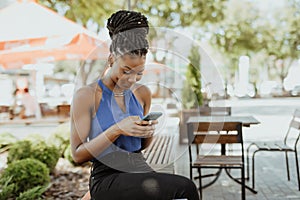 Portrait of young african woman sitting outside on bench reading a text message on her mobile phone