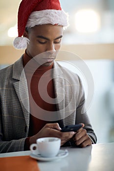 Portrait of young afro-american in the office, texting on cell phone, wearing santa hat
