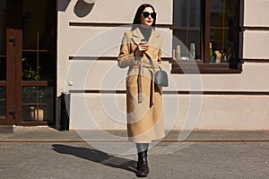 Portrait of young amazing woman standing in front of popular restaurant, wears stylish sunglasses, mustard coat, leather bag and