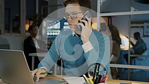 Portrait of young ambitious businessman talking on mobile phone and using laptop in office