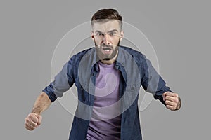 Portrait of young angry man shouting with rage, clenching fists on grey studio background