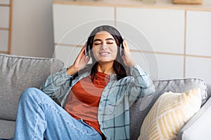 Portrait of young Arab woman in headphones relaxing on couch and listening to music with closed eyes at home