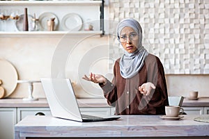 Portrait of a young Arab woman in hijab looking worriedly at the camera and spreading her hands. Sitting at home in the